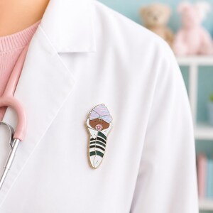 Pode incluir: Um broche de esmalte em tom dourado com um beb&eacute; enrolado em mantas de pele escura, usando um gorro &agrave;s riscas roxo claro e branco. O beb&eacute; tem uma pequena chupeta cor-de-rosa na boca e est&aacute; enrolado numa manta branca com riscas horizontais verde-escuras.