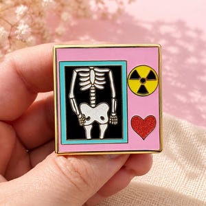 Puede incluir: Un pin de esmalte cuadrado con un borde de metal dorado. El fondo es rosa claro, con un marco rectangular central de interior negro que muestra un torso esquelético humano blanco. A la derecha, un símbolo de radiación amarillo y un corazón rojo brillante.