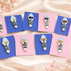 Pode incluir: Uma cole&ccedil;&atilde;o de pins de esmalte, cada um apresentando um beb&eacute; enrolado em mantas com um gorro azul claro e uma chupeta. Os beb&eacute;s est&atilde;o embrulhados em mantas brancas com riscas verde-escuras, montados em pequenos cart&otilde;es retangulares azuis ou cor-de-rosa.