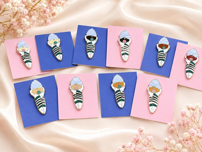 Pode incluir: Uma cole&ccedil;&atilde;o de pins de esmalte, cada um apresentando um beb&eacute; enrolado em mantas com um gorro azul claro e uma chupeta. Os beb&eacute;s est&atilde;o embrulhados em mantas brancas com riscas verde-escuras, montados em pequenos cart&otilde;es retangulares azuis ou cor-de-rosa.
