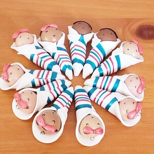 May include: A set of 12 handmade baby dolls, each wrapped in a white and blue striped blanket with pink accents. The dolls have brown skin and are all facing forward.