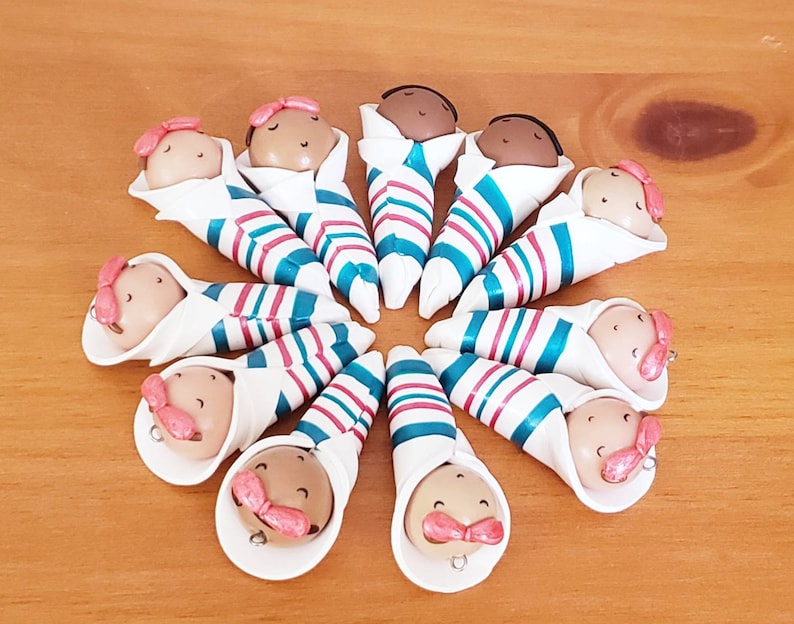 May include: A set of 12 handmade baby dolls, each wrapped in a white and blue striped blanket with pink accents. The dolls have brown skin and are all facing forward.