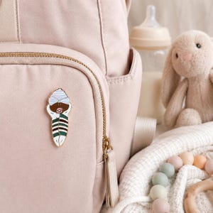 Pode incluir: Um broche de esmalte em tom dourado com um beb&eacute; enrolado em mantas de pele escura, usando um gorro &agrave;s riscas azul-claro e branco e uma chupeta cor-de-rosa. O beb&eacute; est&aacute; embrulhado numa manta &agrave;s riscas verdes, brancas e castanhas, preso a uma mochila rosa.