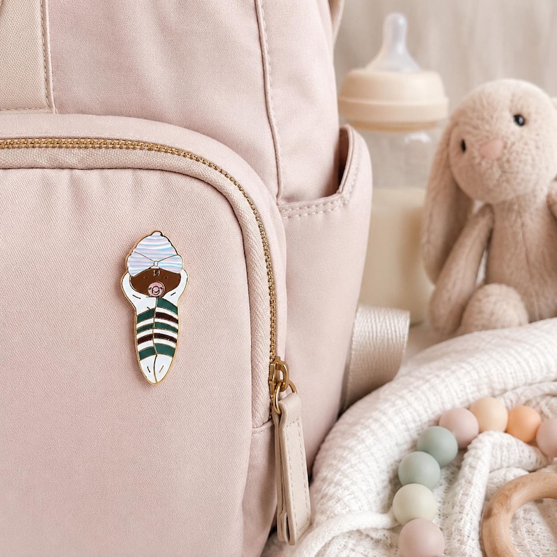 Pode incluir: Um broche de esmalte em tom dourado com um beb&eacute; enrolado em mantas de pele escura, usando um gorro &agrave;s riscas azul-claro e branco e uma chupeta cor-de-rosa. O beb&eacute; est&aacute; embrulhado numa manta &agrave;s riscas verdes, brancas e castanhas, preso a uma mochila rosa.