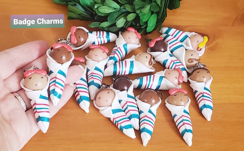 May include: A collection of 14 handmade baby doll charms, each wrapped in a white, teal, and pink striped blanket. The charms are made of polymer clay and have painted facial features. The charms are attached to key rings, making them perfect for adding a touch of whimsy to bags, backpacks, or keys.