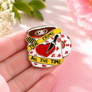 May include: A metal enamel pin shaped like a coffee mug with a white base, red heart, and black knife graphic. A yellow caution tape banner wraps around the mug with the black text TRUE CRIME ALL THE TIME. The mug features red splatter accents.