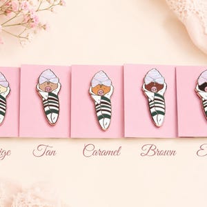 Pode incluir: Cinco pins de esmalte apresentando um beb&eacute; enrolado com um gorro roxo claro e uma chupeta rosa, cada um com um tom de pele diferente rotulado como Beige, Tan, Caramel, Brown e Ebony. Os beb&eacute;s est&atilde;o enrolados em mantas brancas com riscas verde-escuras.