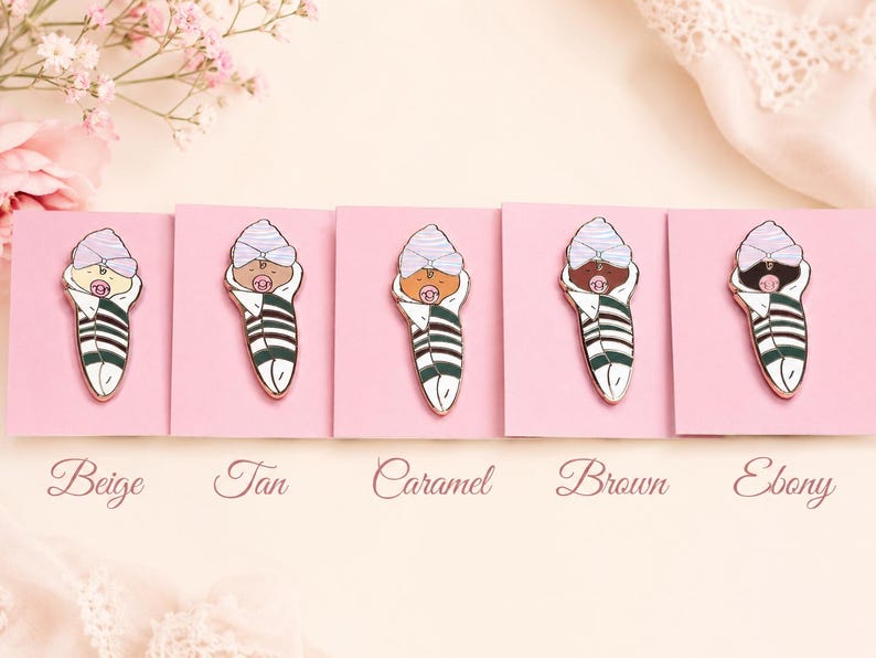 Pode incluir: Cinco pins de esmalte apresentando um beb&eacute; enrolado com um gorro roxo claro e uma chupeta rosa, cada um com um tom de pele diferente rotulado como Beige, Tan, Caramel, Brown e Ebony. Os beb&eacute;s est&atilde;o enrolados em mantas brancas com riscas verde-escuras.