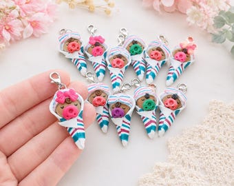 May include: A collection of small polymer clay charms shaped like swaddled babies. Each baby has a tan face, a colorful pacifier, and a striped blanket in shades of teal, magenta, and white. Some feature small bows, and all include a metal lobster clasp.