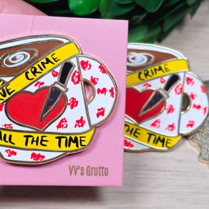 May include: A gold-colored enamel pin featuring a coffee cup with a red heart, a knife, and yellow caution tape that reads "True Crime All The Time".