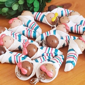 May include: A collection of handmade baby doll keychains. Each doll is wrapped in a white fabric with teal and pink stripes and has a brown head with pink bows.