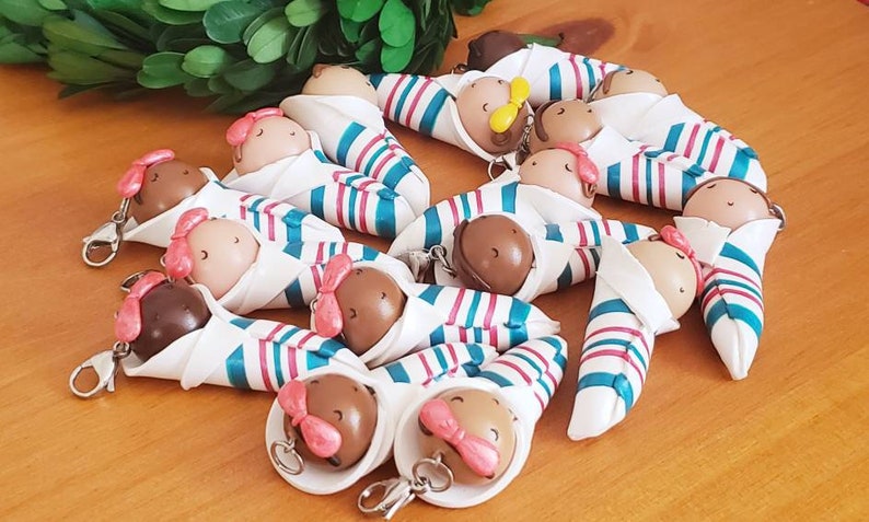 May include: A collection of handmade baby doll keychains. Each doll is wrapped in a white fabric with teal and pink stripes and has a brown head with pink bows.