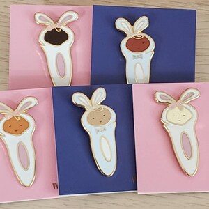 May include: Five enamel pins featuring a cartoon baby wearing a bunny costume. The pins are on pink and blue backgrounds. The babies have different skin tones and are wearing different coloured outfits. The pins are all gold-toned.