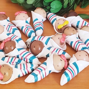 May include: A collection of 14 handmade baby doll keychains. Each doll is wrapped in a white, blue, and pink striped blanket and has a brown face with painted eyes and a pink bow.