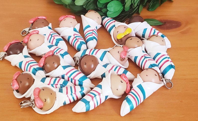 May include: A collection of 14 handmade baby doll keychains. Each doll is wrapped in a white, blue, and pink striped blanket and has a brown face with painted eyes and a pink bow.
