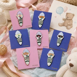 Pode incluir: Sete pins de esmalte com um beb&eacute; enrolado numa manta branca com riscas verde-escuras, usando um gorro &agrave;s riscas azuis ou roxas e uma chupeta. Cada pin est&aacute; montado num cart&atilde;o quadrado cor-de-rosa ou azul com a etiqueta VV's Grotto.