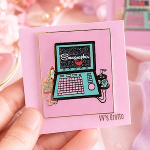 May include: A rectangular enamel pin featuring a light blue ultrasound machine with a black screen displaying the word Sonographer and a red heart. The pin is mounted on a light pink backing card with the text VV's Grotto printed in the corner.