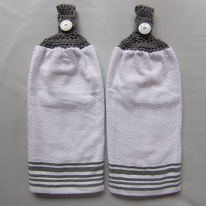 May include: Two white kitchen towels with gray crocheted tops and gray and white striped bottoms. Each towel has a white button on the top.
