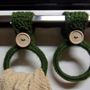 May include: Two handmade green crochet towel holders with wooden buttons. The holders are attached to a metal bar, with a tan towel hanging from one. The crochet work is detailed and the design is simple and functional.