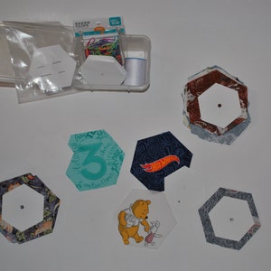 May include: A collection of fabric hexagons with various designs, including a Winnie the Pooh character, a Hot Wheels logo, and a number "3". The hexagons are arranged on a white surface. There is a container with paper clips and a white hexagon template.