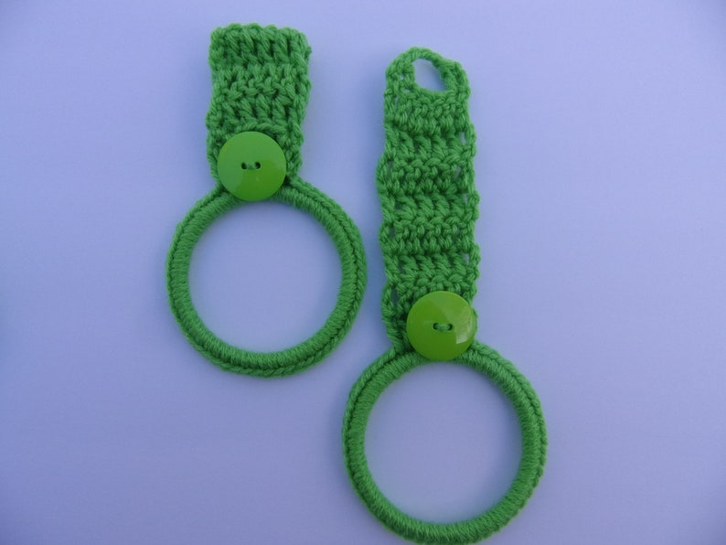 May include: Two green crocheted dish towels with green buttons. The towels are hanging on a white background.