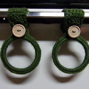 May include: Two green crocheted oven mitts with wooden buttons. The mitts are hanging on a metal oven rack.