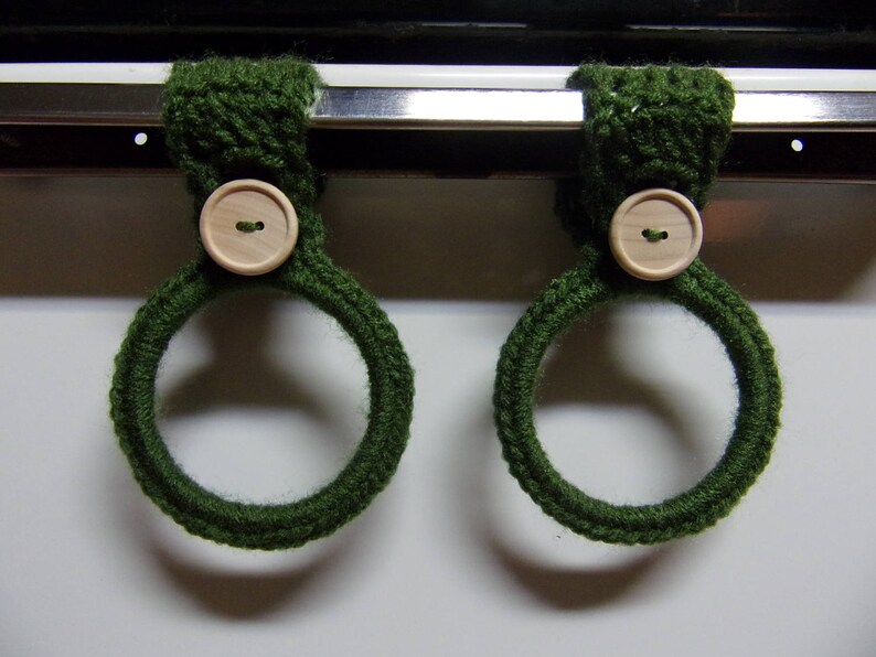 May include: Two green crocheted oven mitts with wooden buttons. The mitts are hanging on a metal oven rack.