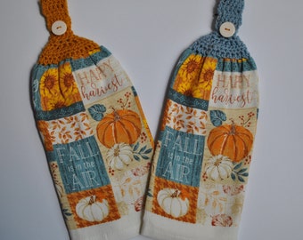 May include: Two kitchen towels with a fall theme. The towels are white with a patchwork design featuring pumpkins, sunflowers, and leaves in shades of orange, yellow, brown, and green. The text "Happy Harvest" and "Fall is in the Air" is printed on the towels. Each towel has a crocheted loop with a wooden button.