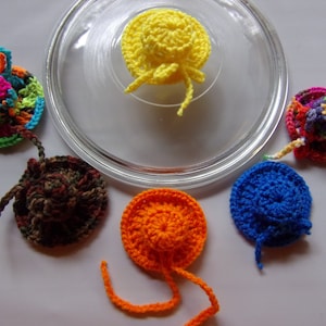 May include: Six crocheted hats in various colors, including yellow, orange, blue, green, and brown. The hats are arranged on a white surface with a glass lid in the background.