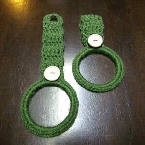 May include: Two green crocheted dishcloths with white buttons. The dishcloths are in the shape of a ring.