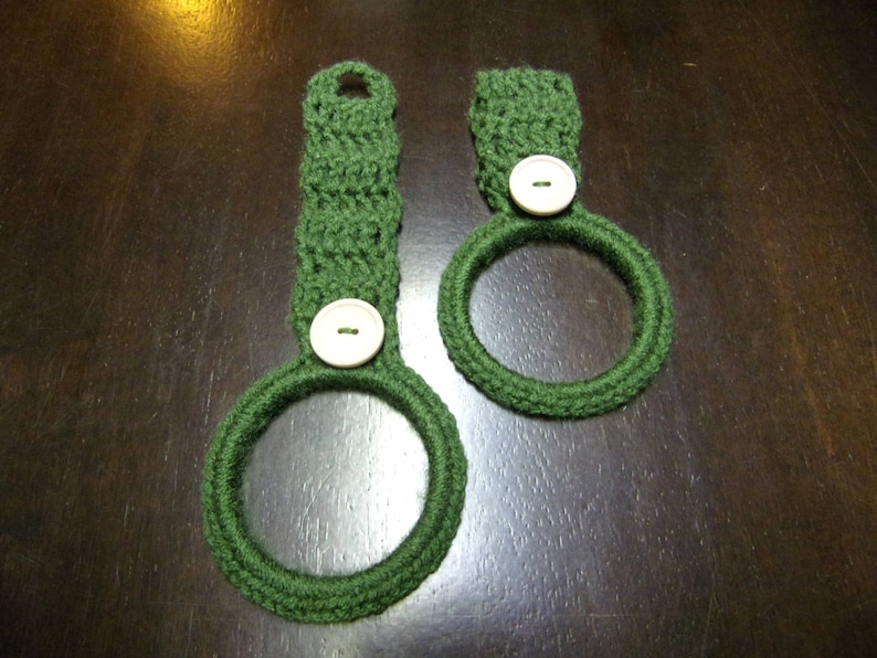 May include: Two green crocheted dishcloths with white buttons. The dishcloths are in the shape of a ring.
