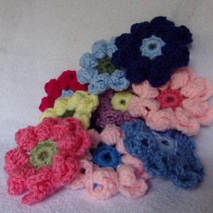 May include: A collection of crocheted flowers in various colors, including pink, blue, yellow, purple, and green. The flowers are arranged in a cluster, with the largest flower in the center.