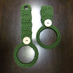 May include: Two green crocheted dishcloths with white buttons. The dishcloths are shaped like rings.