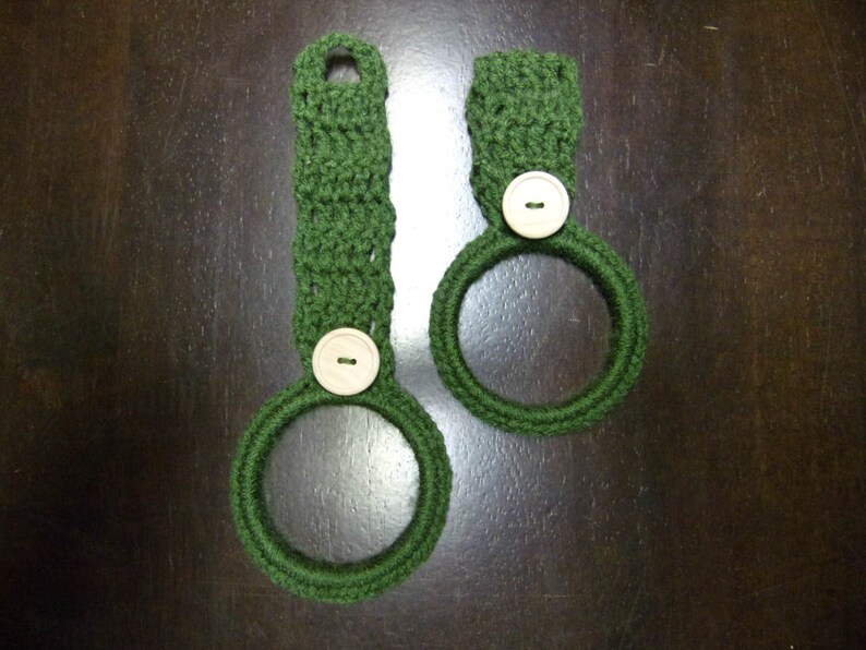May include: Two green crocheted dishcloths with white buttons. The dishcloths are shaped like rings.