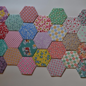 May include: A collection of colorful, hexagonal fabric patches arranged in a honeycomb pattern. Each patch features a unique floral or geometric print in shades of red, pink, blue, green, and yellow. The patches are likely part of a quilt or craft project.