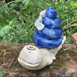 Könnte beinhalten: Eine Keramik-Schneckenfigur mit einem glatten, cremeweißen Körper, zwei kleinen Augen und einem breiten, lächelnden Mund. Das Schneckenhaus ist eine glänzende, tiefblaue Spirale, die mit mehreren kleinen, weißen, geformten Blüten verziert ist.