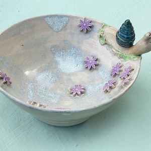 Op de afbeelding: Een keramische kom met een lichtgrijze, gespikkelde glazuurlaag en diverse kleine, verhoogde paarse bloemapplicaties aan de binnenzijde. Een gebeeldhouwde slak met een donkerblauw, spiraalvormig huisje is aan de rand bevestigd.
