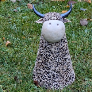 May include: A ceramic Highland cow sculpture with a cream-colored head, black horns, and a shaggy, textured body. The sculpture is set against a backdrop of green grass and fallen leaves, showcasing its rustic charm.