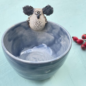 Può includere: Una ciotola in ceramica con una smaltatura lucida marmorizzata blu e grigia. Una piccola creatura scolpita di colore chiaro, con orecchie scure testurizzate e un motivo a tratteggio incrociato sulla pancia, è fissata al bordo interno.