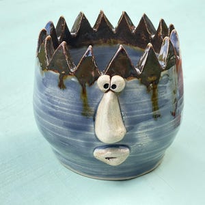 Może przedstawiać: Ceramiczna doniczka z niebieskim szkliwem i ząbkowanym, ciemnobrązowym brzegiem. Naczynie posiada wyrzeźbioną twarz z dwojgiem okrągłych oczu, wydatnym nosem i małymi ustami, co nadaje mu wygląd przypominający postać na przedniej stronie.