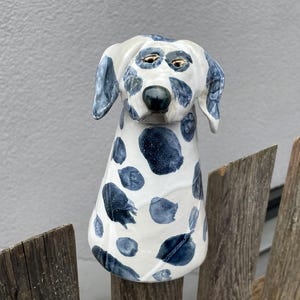 Op de afbeelding: Een keramieken hondenbeeldje met een witte basis en marineblauwe vlekken. De hond heeft een zwarte neus, goudkleurige ogen en flaporen. Het beeldje staat voor een houten hek.