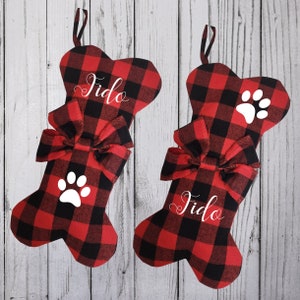 Op de afbeelding: Twee rode en zwarte kerstsokken in buffalo plaid-patroon in de vorm van een hondenbot met witte poot afdrukken en de naam "Fido" geborduurd.