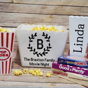 May include: Three white popcorn containers with black lettering. The largest container says "The Braxton Family Movie Night" with a wreath and the letter "B" in the centre. The medium container says "Linda". The smallest container says "POP CORN" with red and white stripes. There are also several candy bars stacked on top of each other.