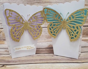 Puede incluir: Dos cajas de regalo blancas con recortes de mariposas. Las mariposas son de purpurina dorada con detalles en morado y turquesa. Una caja tiene una pancarta blanca con el texto "Happy 1st Birthday Wynter".