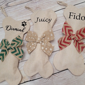 May include: Three beige bone-shaped Christmas stockings with different names and bows. The stockings are decorated with green, brown, and red bows. The names on the stockings are "Diamond", "Juicy", and "Fido".
