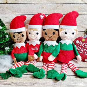 Può includere: Quattro bambole di elfo di peluche con abiti rossi e verdi, cappelli rossi e bordi bianchi. Gli elfi sono seduti su una superficie di legno con un albero di Natale verde e un ornamento rosso e dorato con il testo "Buon Natale".