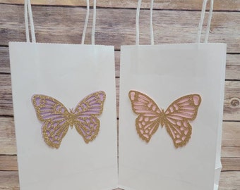 Puede incluir: Dos bolsas de regalo de papel blanco con diseños de mariposas doradas con purpurina. Una mariposa es morada y la otra es rosa.