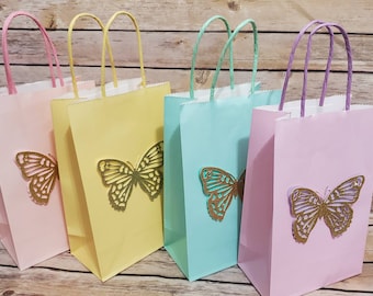 Puede incluir: Cuatro bolsas de regalo de papel de color pastel con recortes de mariposas doradas. Las bolsas son rosa, amarilla, azul claro y lavanda.