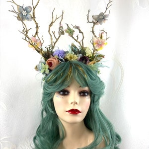May include: A green wig with a floral and twig crown. The crown features delicate flowers in shades of pink, blue, and white, and gold accents.