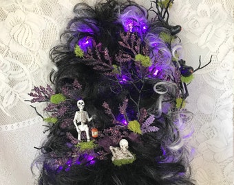 May include: A Halloween-themed wig with black and gray hair, decorated with purple lights, miniature skeletons, and sprigs of purple and green foliage. The wig is styled with a voluminous, textured appearance, perfect for a costume.